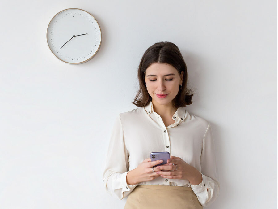 Eine Frau steht unter einer Uhr und tippt auf ihr Smartphone. Sie hat Probleme mit dem Zeitmanagagement.