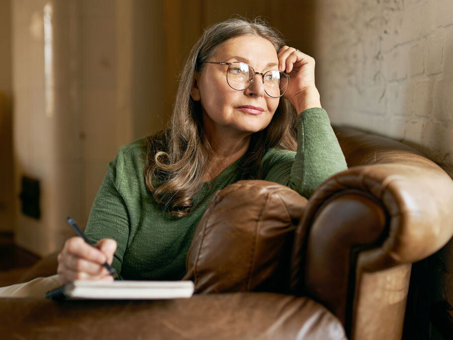 Eine Frau mit grauen Haaren und grünem Pullover sitzt auf einem Sofa und schreibt.