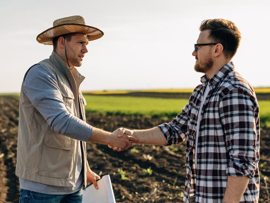 Zwei Männer stehen vor einem Feld und schütteln sich die Hände. Der eine hält einen Landpachtvertrag in den Händen.