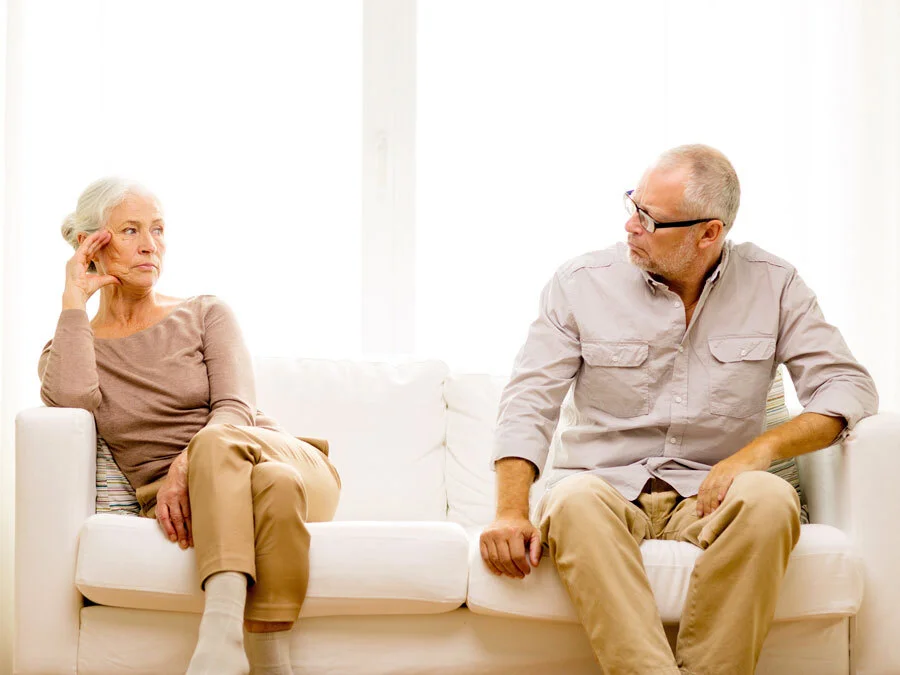 Eine Frau und ein Mann mit grauem Haar sitzen auf einem Sofa und blicken sich feindseelig an.