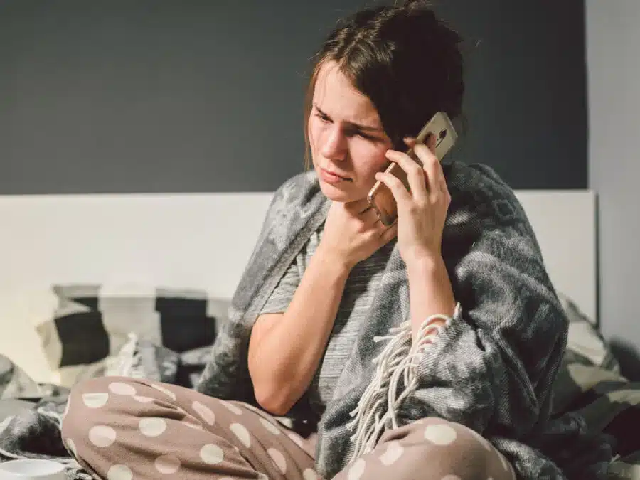 Eine Frau mit Halsschmerzen sitzt auf dem Bett und telefoniert.